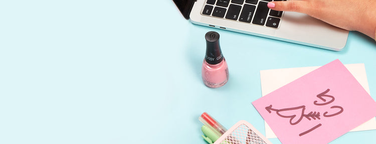 Hand typing on a laptop with pink nail lacquer, a bottle of pink nail lacquer, and stationery items on a light blue background.