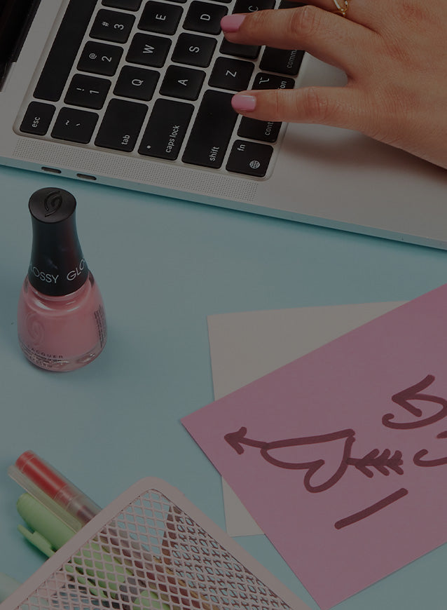 Person typing on a laptop with pink nail lacquer and stationery items on a light blue surface