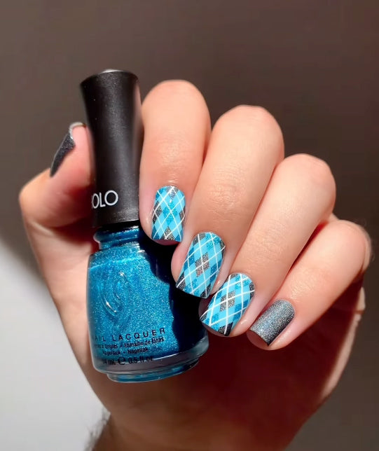 Hand holding a blue glitter nail polish bottle, showing a manicure with blue argyle plaid nails and one silver glitter accent nail.