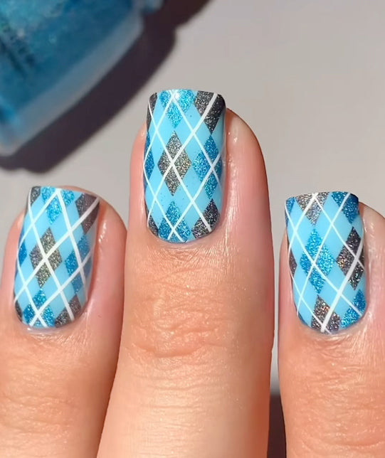 Close-up of fingernails with a blue argyle plaid design featuring light blue, turquoise, silver glitter, and white crisscross lines.
